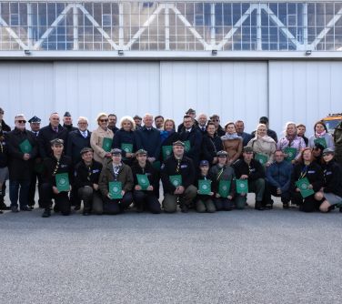 grupa osób pozuje do zdjęcie na płycie lotniska, za nimi hangary, niektórzy trzymają w dłoniach zielone teczki