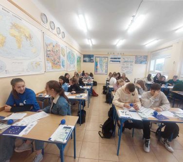 młodzież siedzi w łakwach szkolnych, na ławkach broszury i gadżety o tematyce Unii Europejskiej
