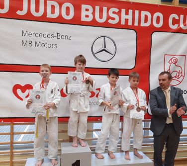 Hala sportowa. Czterech chłopców w kimonach do judo trzymają w dłoniach dyplomy. Po prawej stronie mężczyzna klaszcze