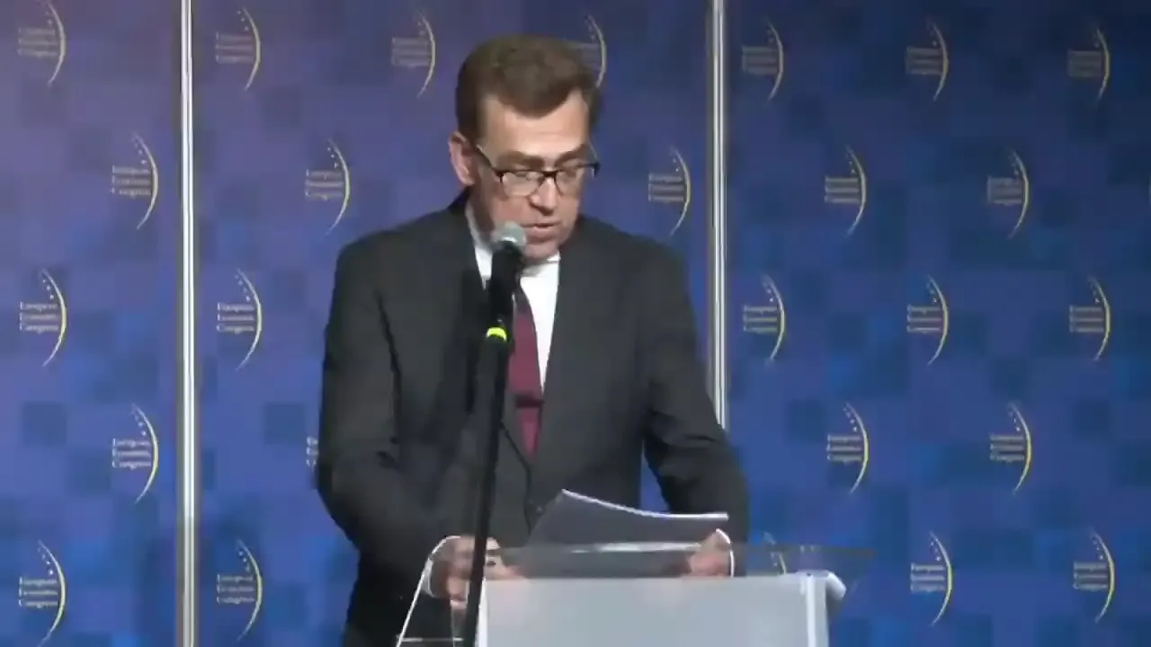 Mężczyzna w garniturze i okularach stoi za mównicą i czyta z kartek przy mikrofonie na tle niebieskiej ścianki z napisem European Congress.