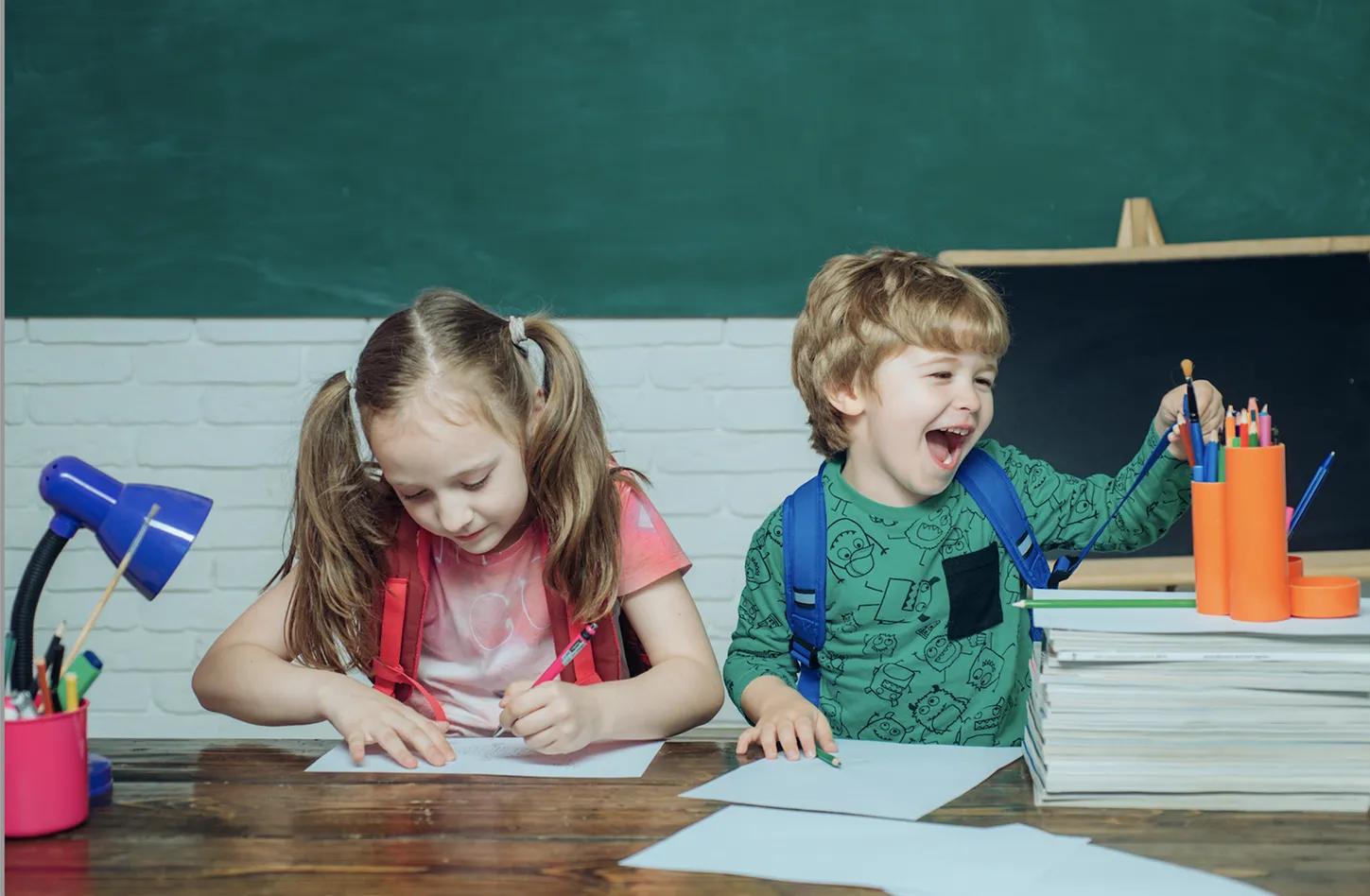 Kliknięcie powiększy obraz. Dwoje dzieci siedzi przy stole z papierem i przyborami do rysowania, jedno pisze, a drugie się śmieje i sięga po kredki.