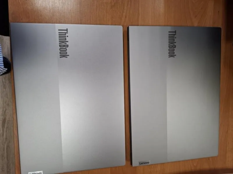 Dwa zamknięte srebrne laptopy Lenovo ThinkBook ułożone obok siebie na drewnianym blacie.