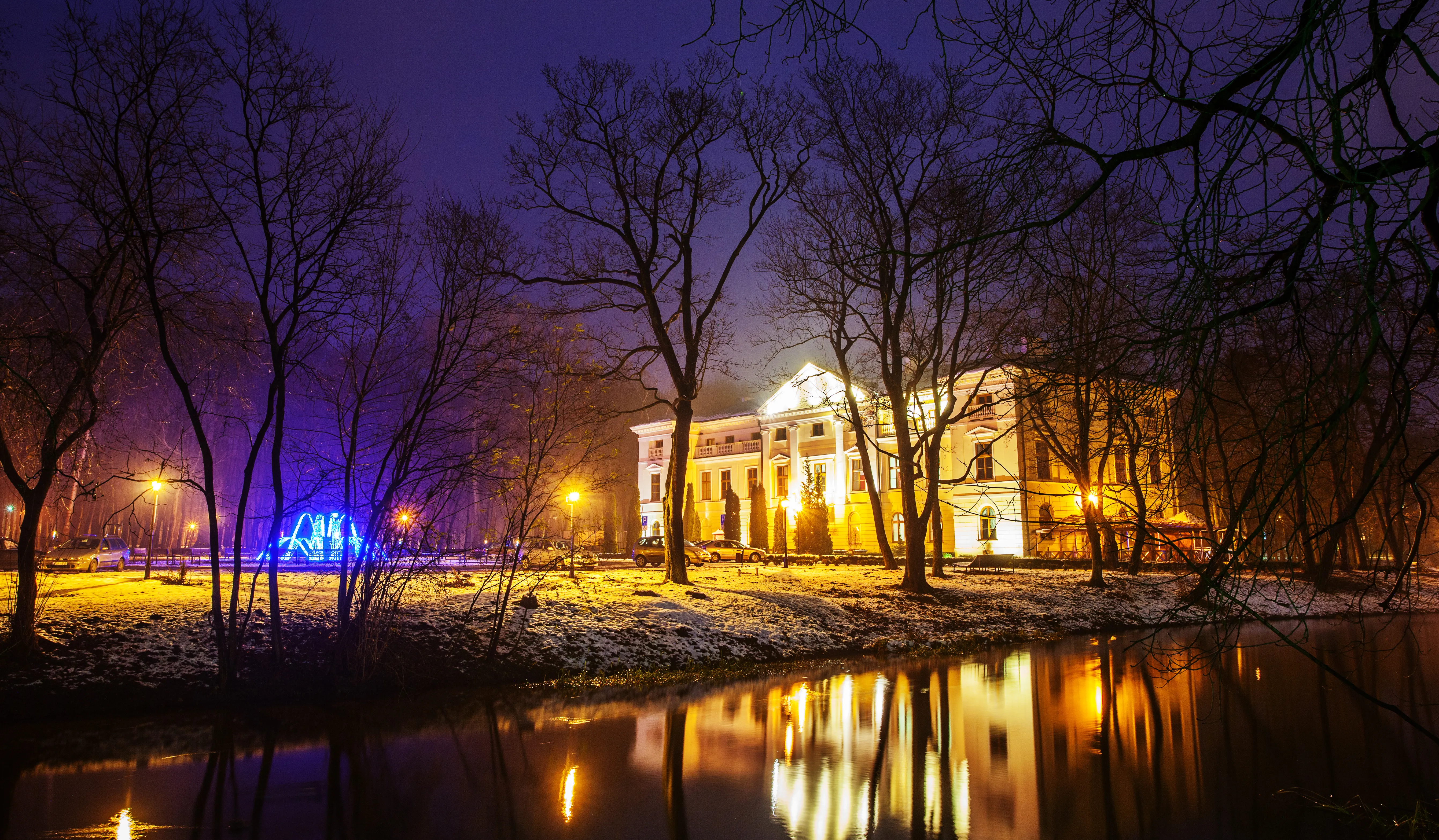Widok nocny na oświetlony pałac w parku z nagimi drzewami i niewielką ilością śniegu na ziemi. Przed pałacem stoi kilka samochodów, w tle niebieskie światełka dekoracyjne. Woda odbija światła budynku i latarni.