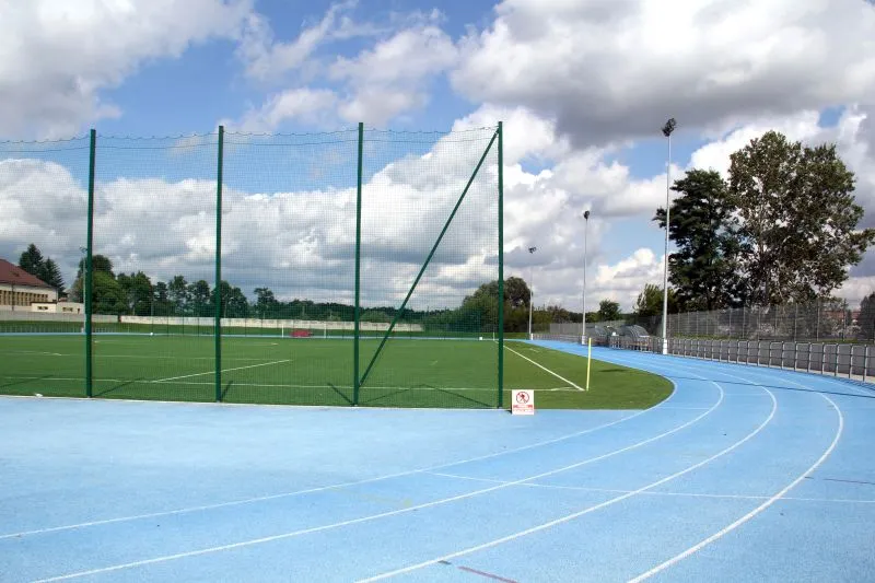 Zielone boisko sportowe z siatką dookoła i niebieska bieżnia z liniami torów.