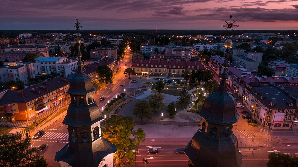 Dwie wieże kościoła w pierwszym planie po obu stronach kadru. Przed nimi oświetlony plac miejski z drzewami, chodnikami i fontanną. W tle ulice z samochodami, budynki mieszkalne i sklepowe oraz fioletowo-różowe niebo o zmierzchu.
