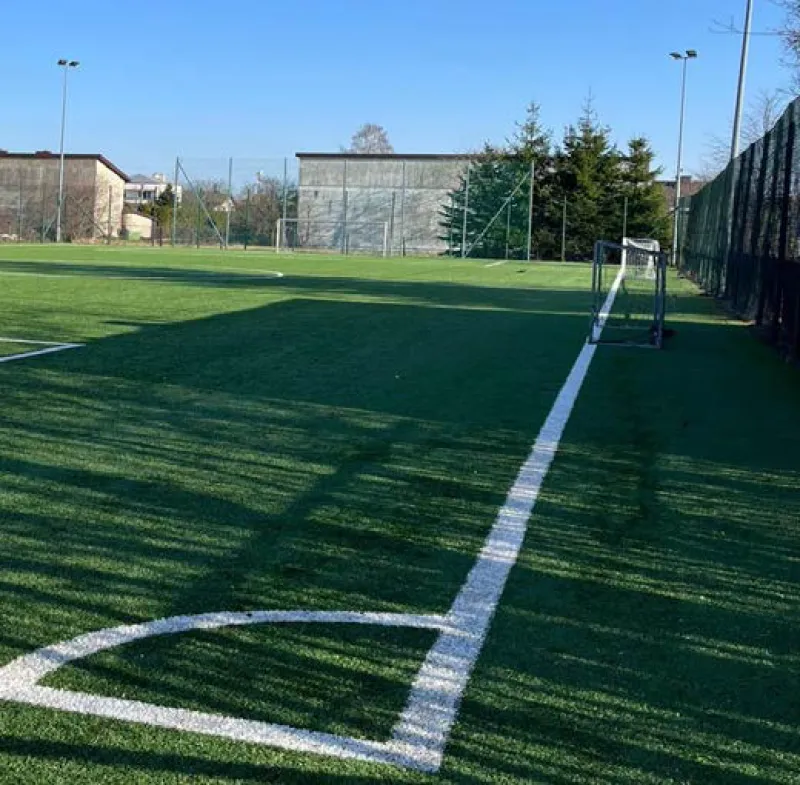 Boisko do piłki nożnej z zieloną nawierzchnią syntetyczną, białymi liniami i bramką treningową po prawej stronie.