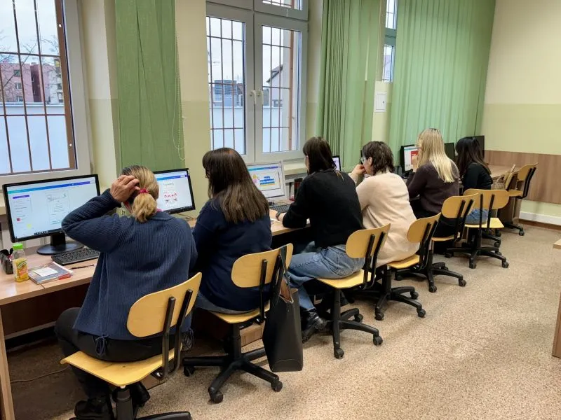 Sześć kobiet siedzi w rzędzie przy biurkach i pracuje na komputerach stacjonarnych w pomieszczeniu z dużymi oknami.