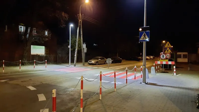 Nocna scena z przejściem dla pieszych oświetlonym latarniami ulicznymi, czerwonobiałe słupki z łańcuchami oraz samochody zaparkowane przy krawężniku.