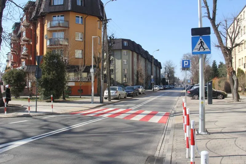 Ulica z oznaczonym przejściem dla pieszych w postaci pasów czerwono-białych, budynki mieszkalne oraz zaparkowane samochody po obu stronach drogi.
