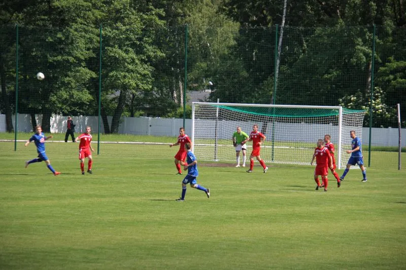 Piłkarze w niebieskich i czerwonych strojach grający na boisku piłkarskim przy bramce z bramkarzem w zielonej koszulce.