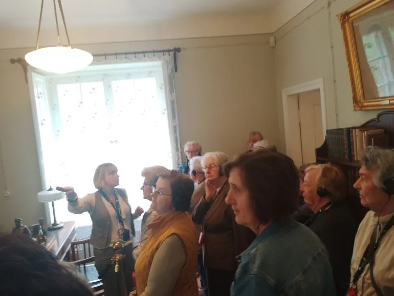 Grupa osób słuchająca kobiety w słuchawkach w pomieszczeniu z regałem na książki i dużym oknem.