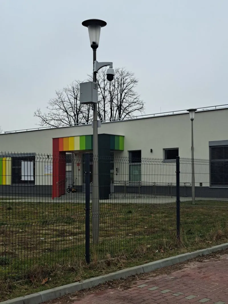 Latarnia z kamerą nad ogrodzeniem na tle budynku z kolorowym wejściem.