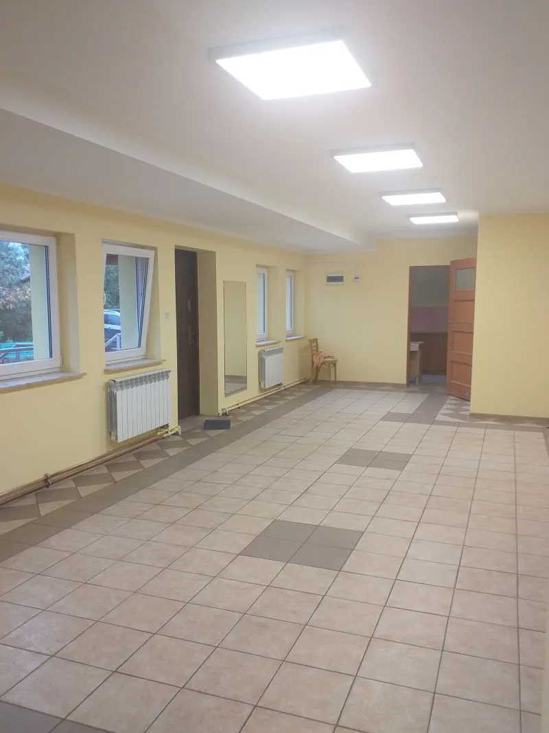 Pusty, jasny pokój z żółtymi ścianami, podłogą wyłożoną kwadratowymi kafelkami, czterema oknami, trzema grzejnikami, lustrem na ścianie i krzesłem przy otwartych drzwiach.