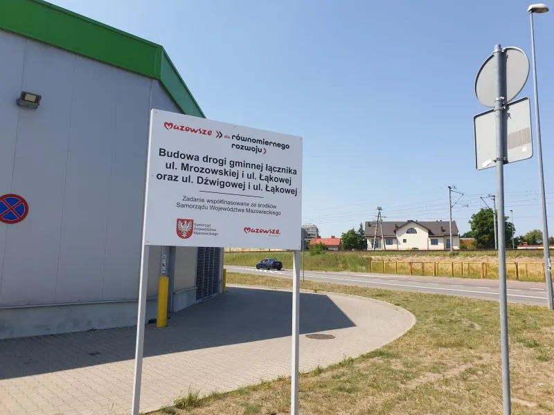 Tablica informacyjna o budowie drogi gminnej łączącej ul. Mrozowską, ul. Łąkową i ul. Dźwigową przy budynku z zielonym dachem.