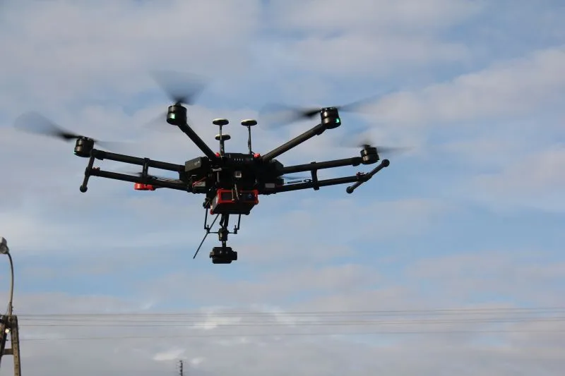 Sześciośmigłowy czarny dron z zamocowaną kamerą w locie na tle nieba.