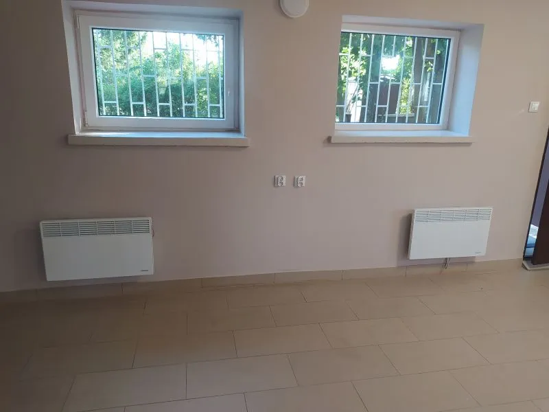 Dwa okna z kratami nad dwoma białymi grzejnikami zamontowanymi na jasnej ścianie i beżowa podłoga z płytek.