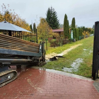 Maszyna budowlana z rurami na gąsienicach stoi przy wyjeździe na trawiasty, częściowo błotnisty teren z ogrodzeniem i drzewami.