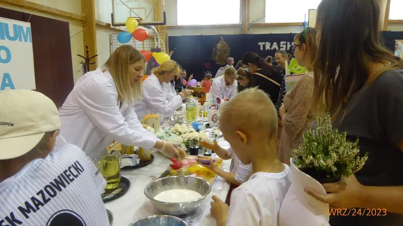 Dorośli w białych fartuchach podają jedzenie dzieciom i dorosłym przy stole z naczyniami i kwiatami.