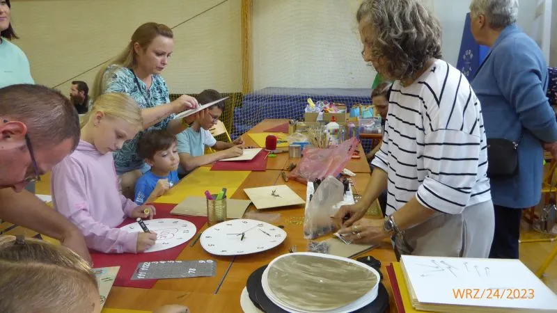 Grupa dzieci i dorosłych siedzi przy stole i wykonuje rękodzieło z wykorzystaniem papieru i materiałów plastycznych.