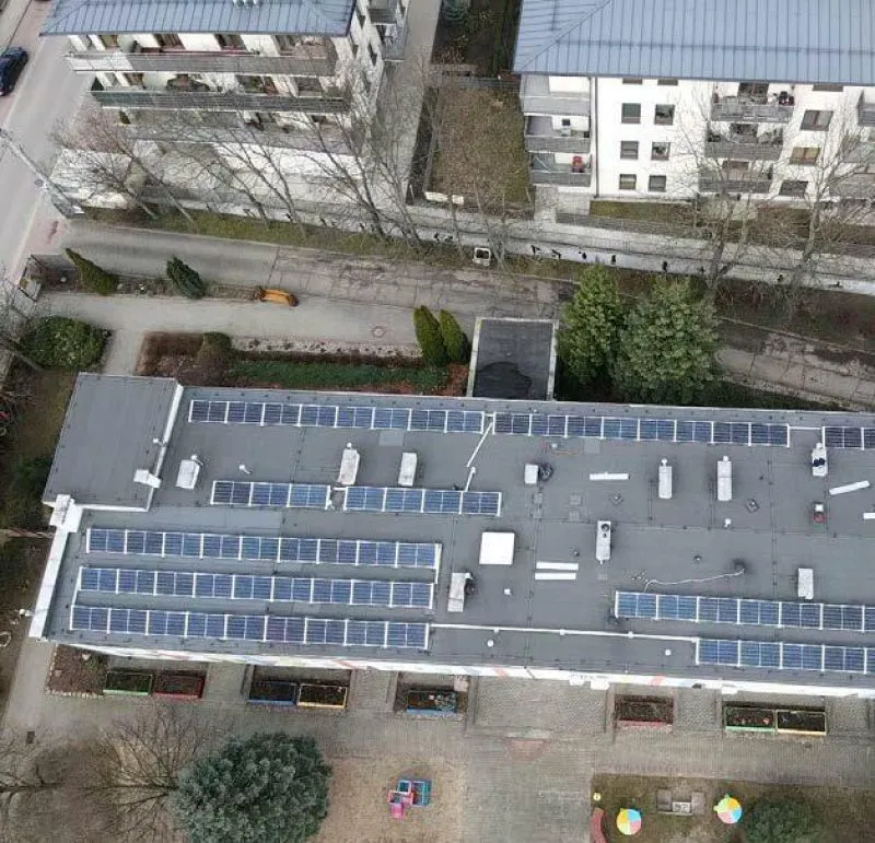 Dach budynku mieszkalnego pokryty panelami słonecznymi, otoczony innymi budynkami i elementami zieleni.