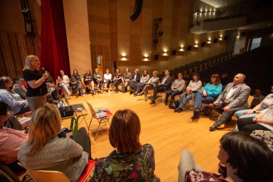 Grupa ludzi siedząca na krzesłach ustawionych w okrąg, jedna kobieta stoi i przemawia przez mikrofon w sali konferencyjnej.