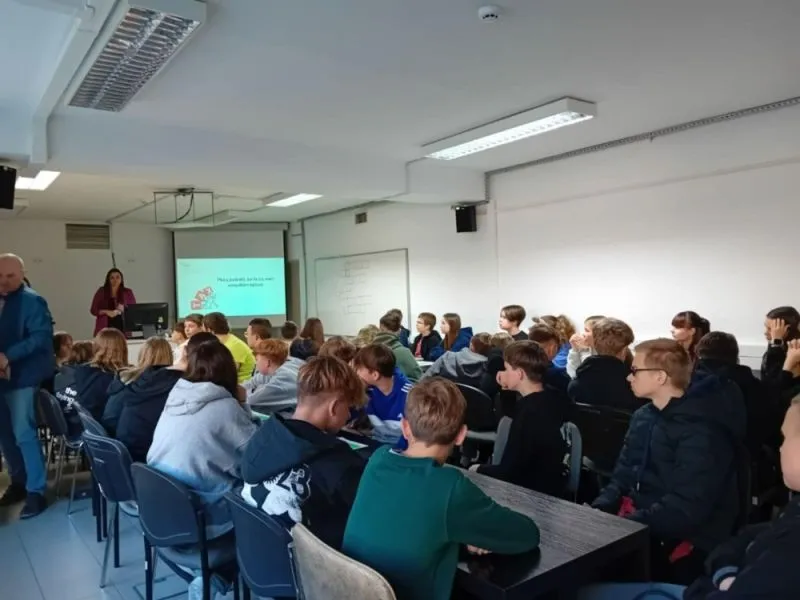 Grupa uczniów siedzi przy stolikach w sali lekcyjnej, a nauczycielka stoi przy projektorze wyświetlającym prezentację.