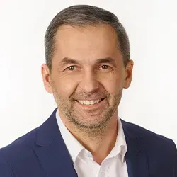 Mężczyzna w średnim wieku ubrany w granatową marynarkę i białą koszulę, uśmiechający się na białym tle.