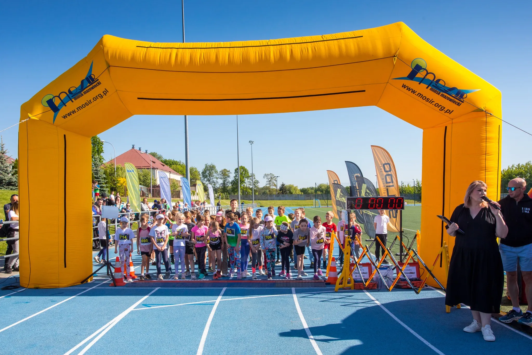 Dzieci ustawione na linii startu biegu pod żółtą, nadmuchiwaną bramą z logo mosir.org.pl, obok kobieta z mikrofonem.