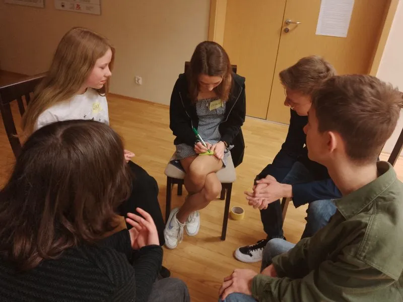 Pięcioro nastolatków siedzi na krzesłach w kręgu, jedna dziewczyna pisze coś na kartce.