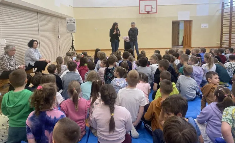 Grupa dzieci siedzi na sali gimnastycznej, słuchając dwójki dorosłych przemawiających na tle kosza do koszykówki.