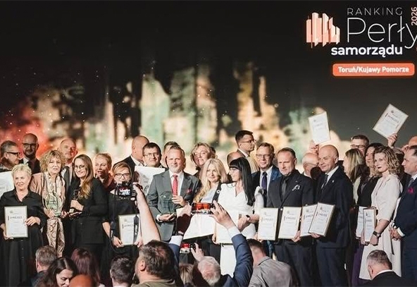 Grupa około stoi na scenie w ubraniach formalnych. Kilka osób trzyma ramki z dyplomami, inni trzymają statuetki. Przed sceną stoją fotografowie i osoby robiące zdjęcia smartfonami. Tło sceny jest ciemne z rozmytym motywem miasta i widocznym logo oraz napisem w prawym górnym rogu.