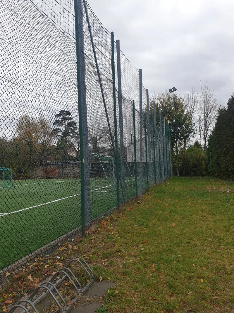Siatkowy ogrodzenie otaczające boisko piłkarskie z widoczną bramką oraz stojakiem na rowery na trawiastym terenie.