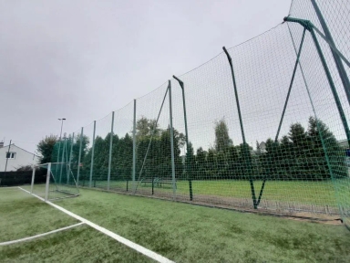 Bramka piłkarska na sztucznej nawierzchni otoczona wysoką siatką ochronną.