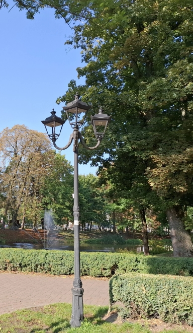 Stary czarny latarnik z trzema lampami stoi na tle zielonych drzew i krzewów w parku przy jasnym, bezchmurnym niebie. W tle widoczna jest fontanna i niewielki staw.