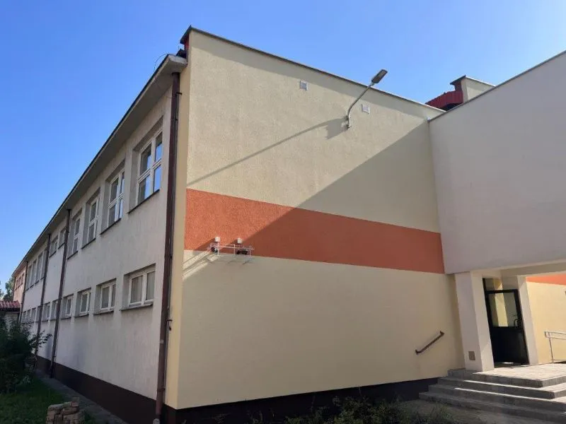 Budynek z dwoma rzędami okien, ściana z poziomym pomarańczowym pasem, schody prowadzące do wejścia oraz lampa na ścianie.