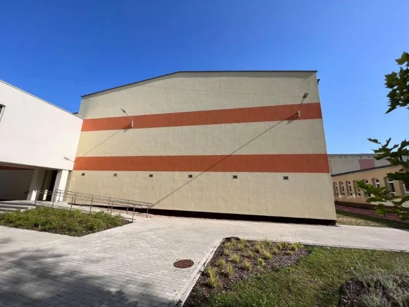 Budynek z jasnymi ścianami ozdobionymi dwoma poziomymi pomarańczowymi pasami oraz podjazdem i chodnikiem z kostki brukowej w otoczeniu zieleni.