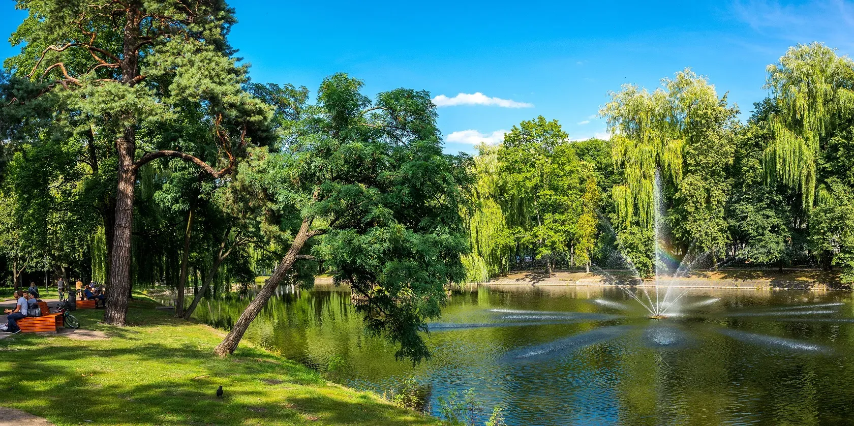 Park ze stawem, fontanną wodną oraz ławkami, przy których siedzą ludzie.