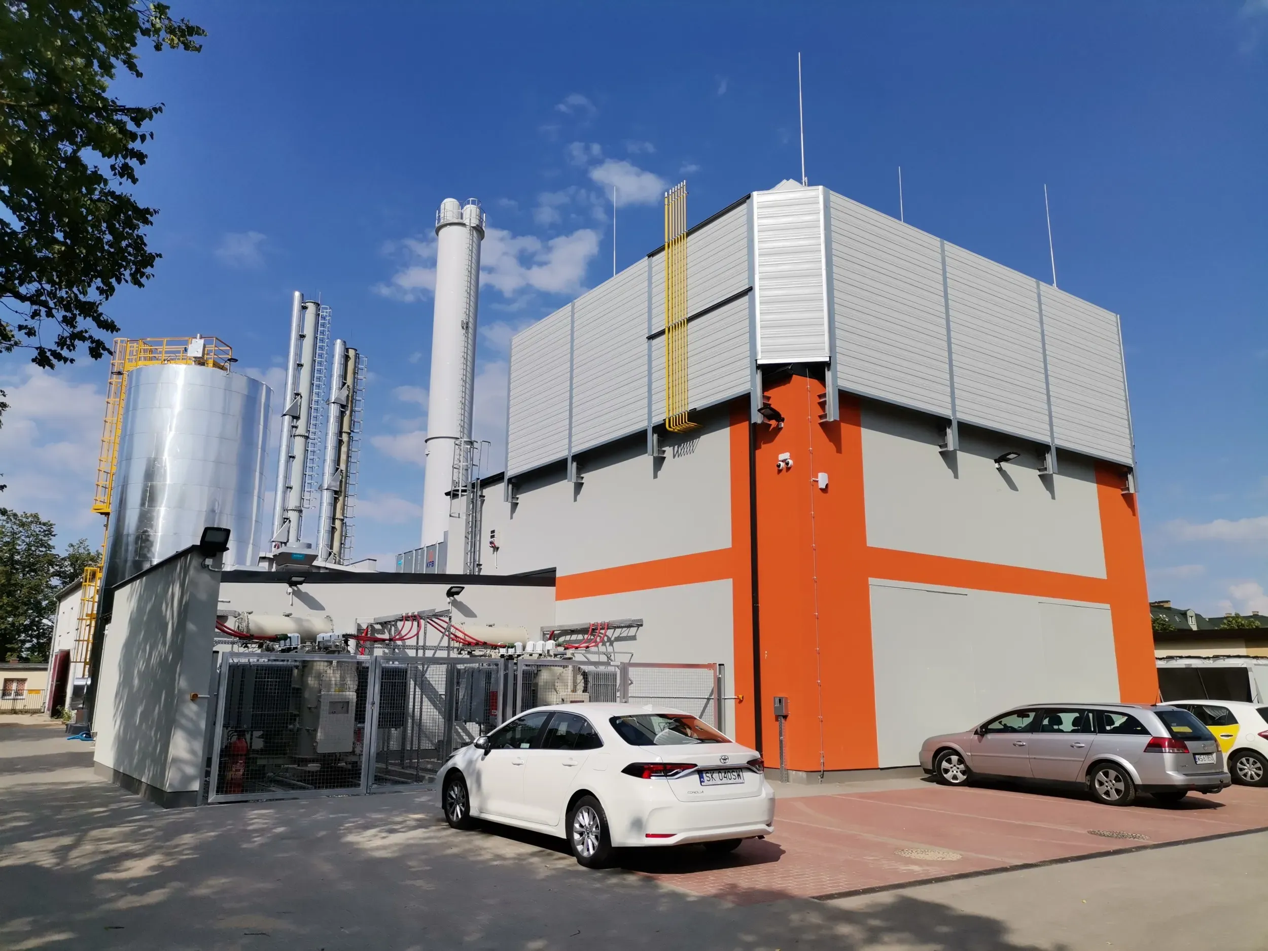 Budynek przemysłowy z instalacjami technicznymi i zaparkowanymi samochodami na pierwszym planie.