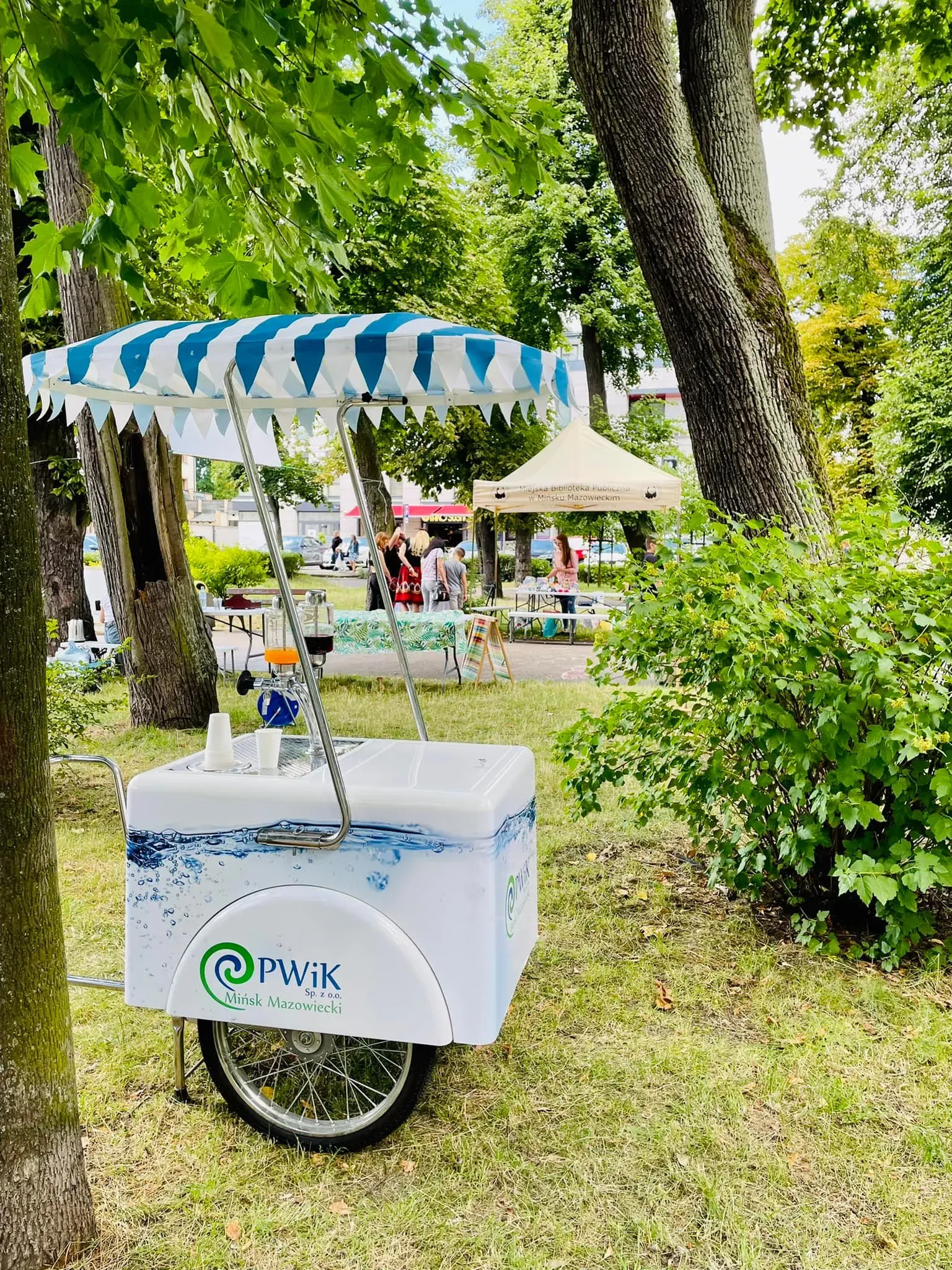 Mobilny wózek z parasolem w biało-niebieskie pasy i logo PWik Mińsk Mazowiecki stoi na trawie między drzewami.