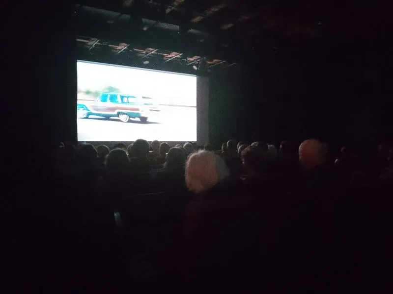 Ciemne kino z widownią oglądającą film na ekranie, na którym widać jadący samochód.