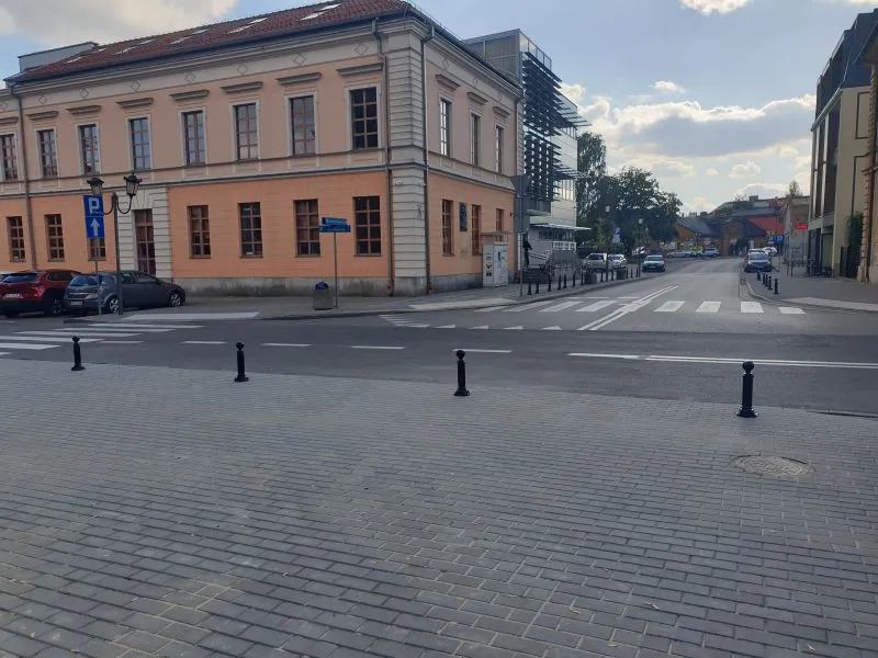 Ulica z przejściem dla pieszych, budynek z beżową elewacją i zaparkowane samochody przy krawężniku.