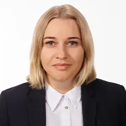 Kobieta o blond włosach ubrana w biały kołnierzyk i ciemną marynarkę na jasnym tle.