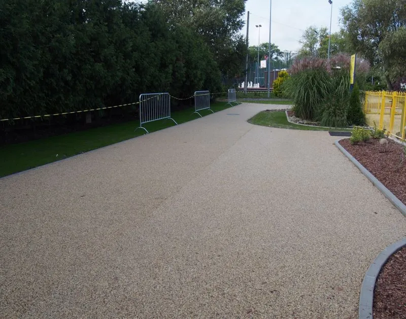 Ścieżka piesza z barierkami po lewej stronie oraz nasadzeniami roślin po prawej stronie i z żółtym ogrodzeniem.