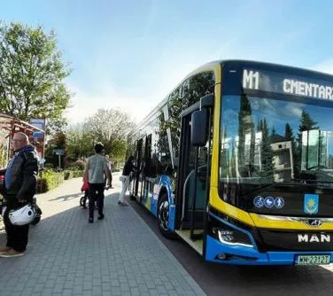 Autobus miejski linii M1 stojący na przystanku, obok niego przechodzi kilka osób.