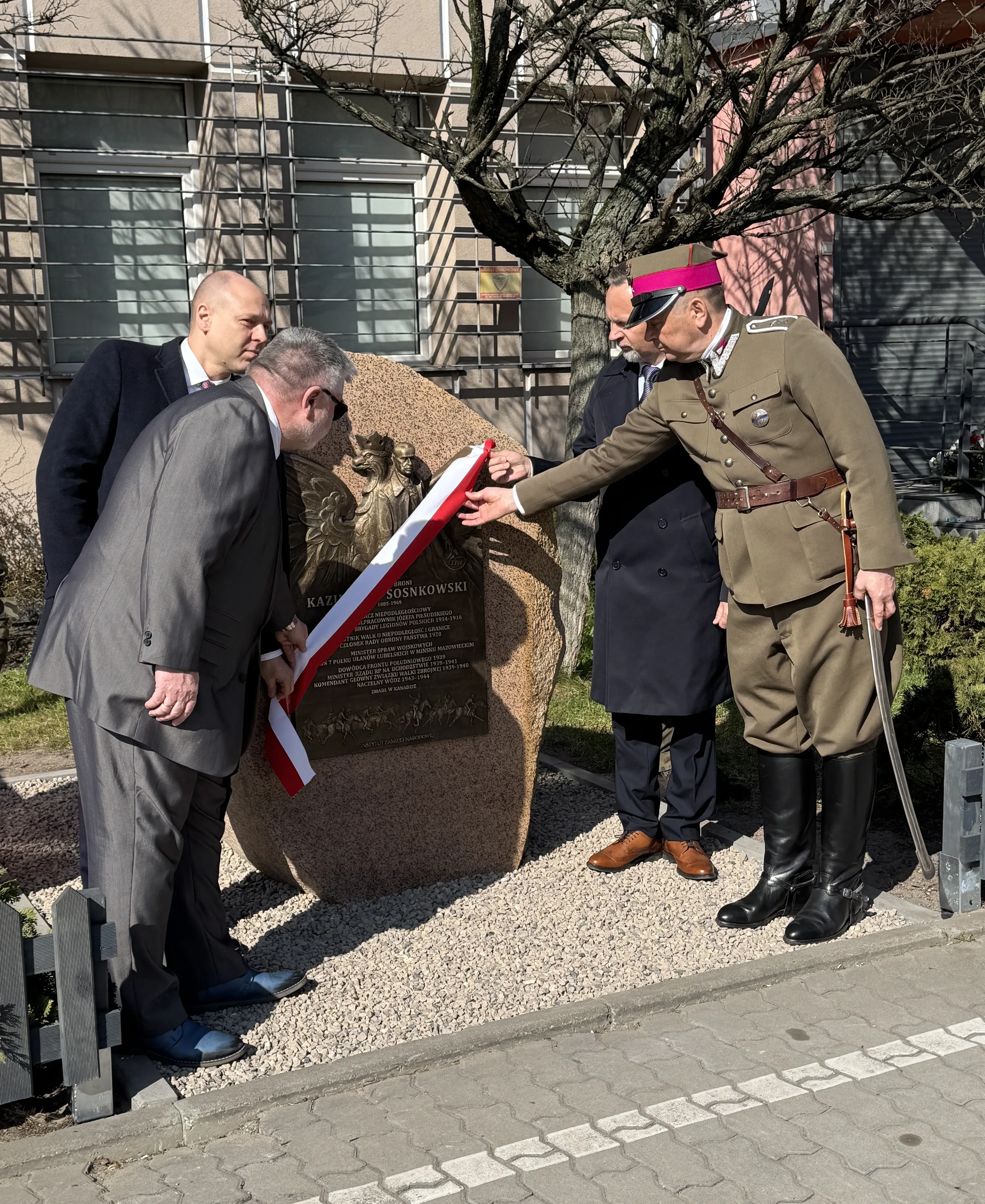 Czterech mężczyzn odsłania kamienną tablicę pamiątkową na zewnątrz w ciągu dnia. Jeden ma historyczny mundur z różowawym paskiem na czapce i szablę, pozostali noszą garnitury i płaszcze. Na tablicy widoczna metalowa płaskorzeźba orła.