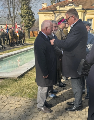 Kilku mężczyzn stoi na zewnątrz. Dwaj mężczyźni na pierwszym planie stoją twarzą do siebie, jeden przypina odznakę drugiemu. W tle są umundurowani żołnierze z pocztami sztandarowymi.