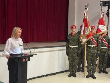 Kobieta stoi przy czarnym pulpicie z mikrofonem przed bordową kotarą. Po prawej stronie stoi czworo żołnierzy w mundurach moro i czerwonych beretach, trzymających flagi z symbolami. Wnętrze jasne, podłoga jasna.
