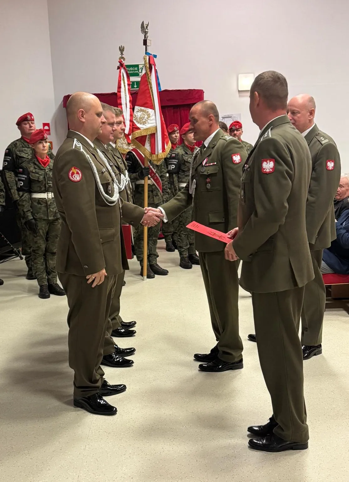 Grupa ośmiu żołnierzy w mundurach przystępuje do ceremonii wewnątrz budynku. Trzech żołnierzy stoi naprzeciwko czterech innych, dwóch z nich podaje sobie ręce. W tle znajduje się pięć osób w mundurach z czerwonymi beretami i patrolową flagą. Ściany są białe, a na podłodze jasny kolor.