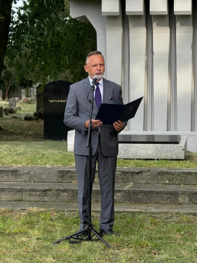 Mężczyzna w garniturze przemawia przy mikrofonie na zewnątrz na tle pomnika i zieleni cmentarnej.