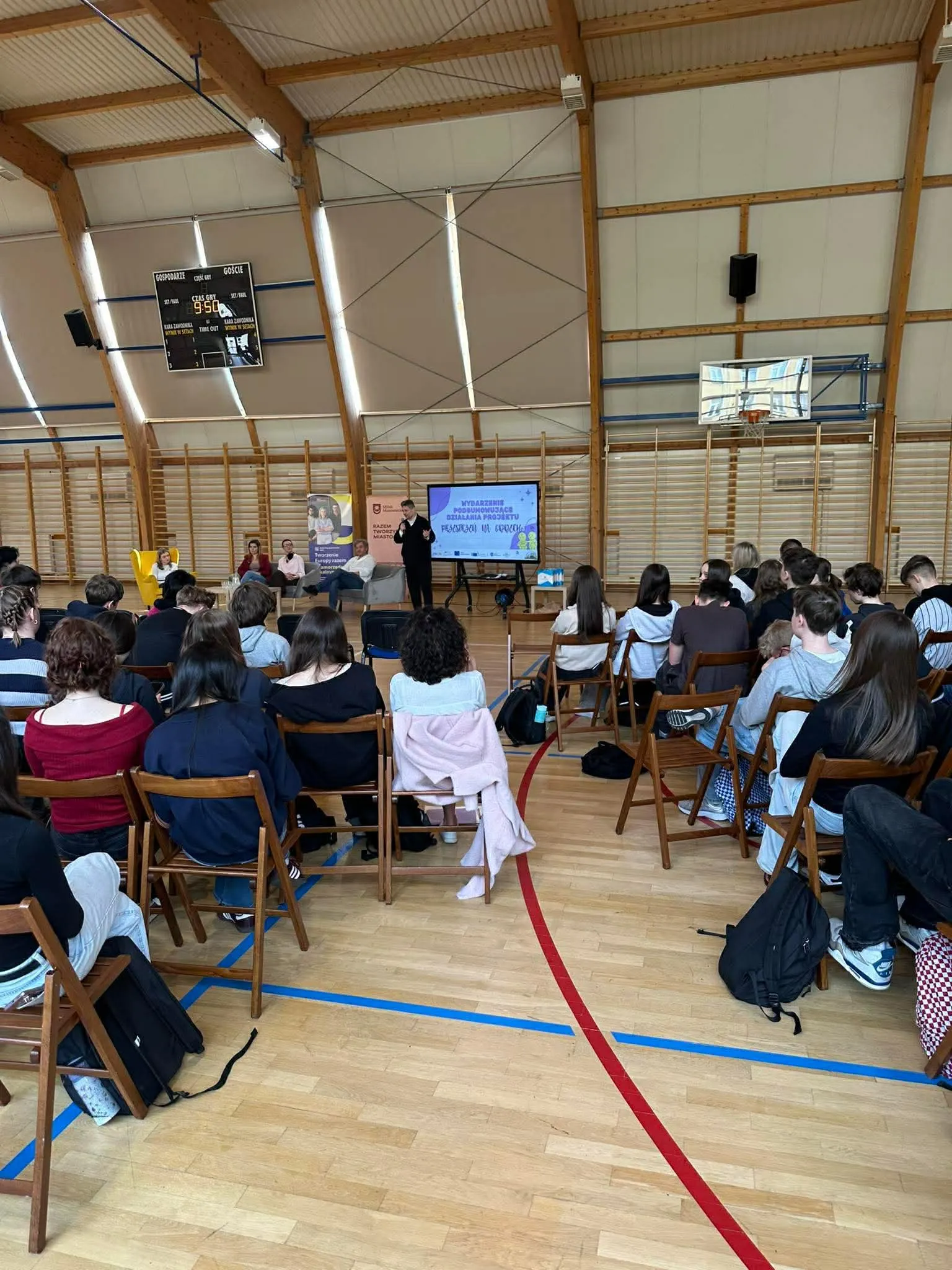 Sala gimnastyczna z drewnianą podłogą i widocznymi czerwonym i niebieskim oznaczeniami boiska. W rzędach składanych krzeseł siedzi grupa uczniów zwróconych przodem do sceny, gdzie stoi prowadzący przy dużym ekranie i kilkoro panelistów na fotelach; w tle tablica wyników i zawieszony kosz.